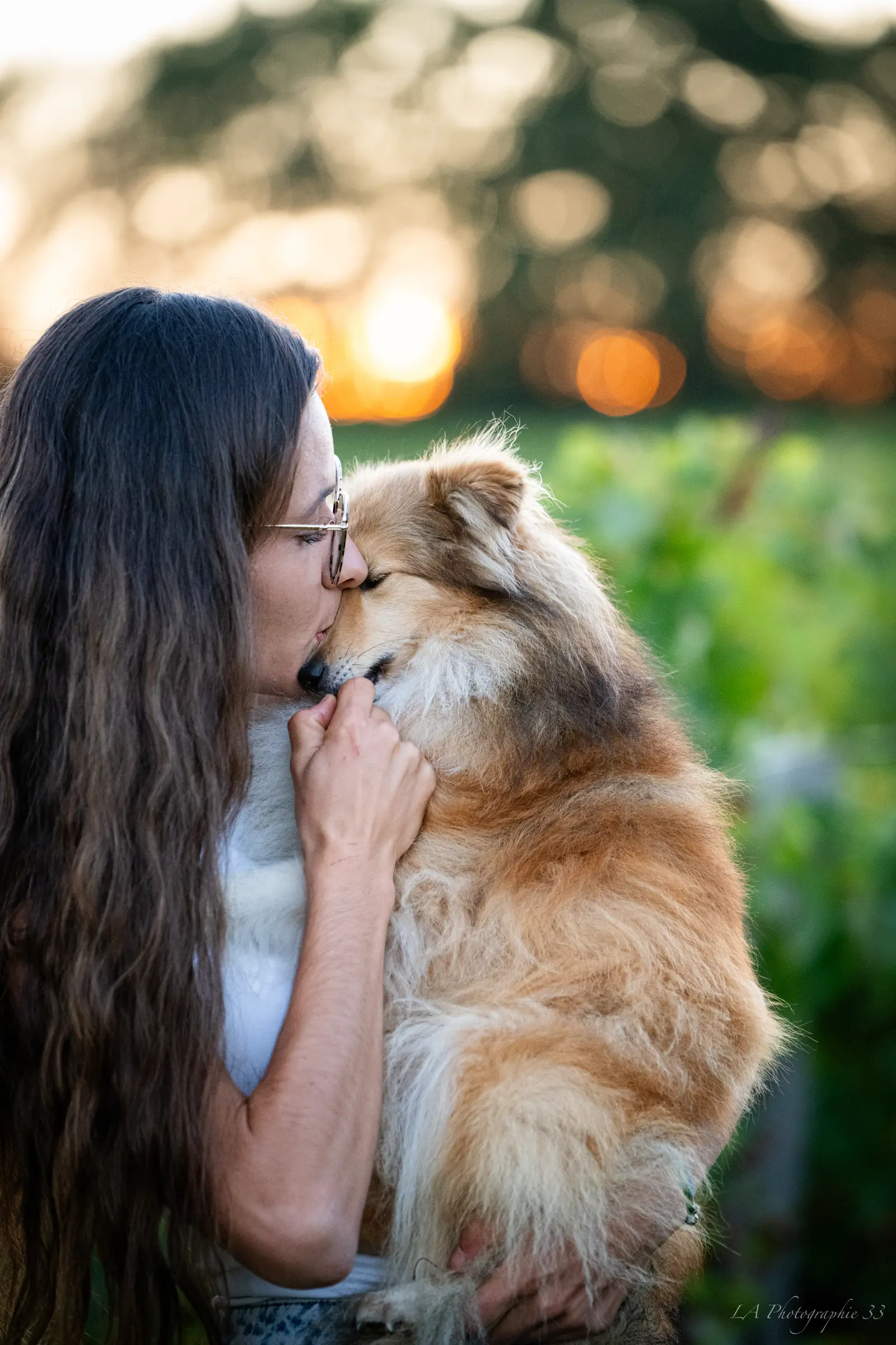 Jeune femme assise dans l'herbe au bord de l'eau avec son chien, éclairés par la lumière dorée du soleil couchant