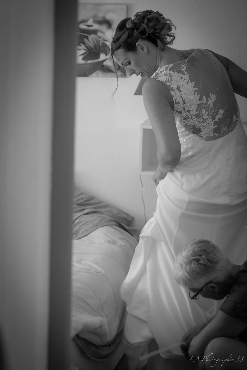 Photo en noir et blanc d'une mariée de dos dont on ajuste la robe en dentelle