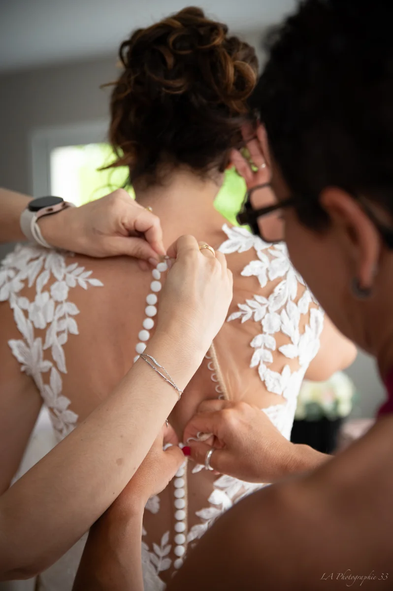 Mariée de dos se faisant boutonner sa robe en dentelle par deux proches