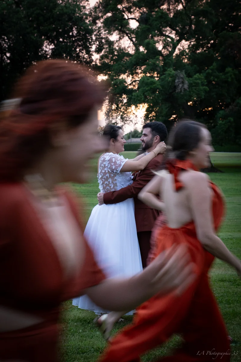 Mariés dansant enlacés au coucher du soleil, demoiselles d'honneur en robes rouges en mouvement au premier plan