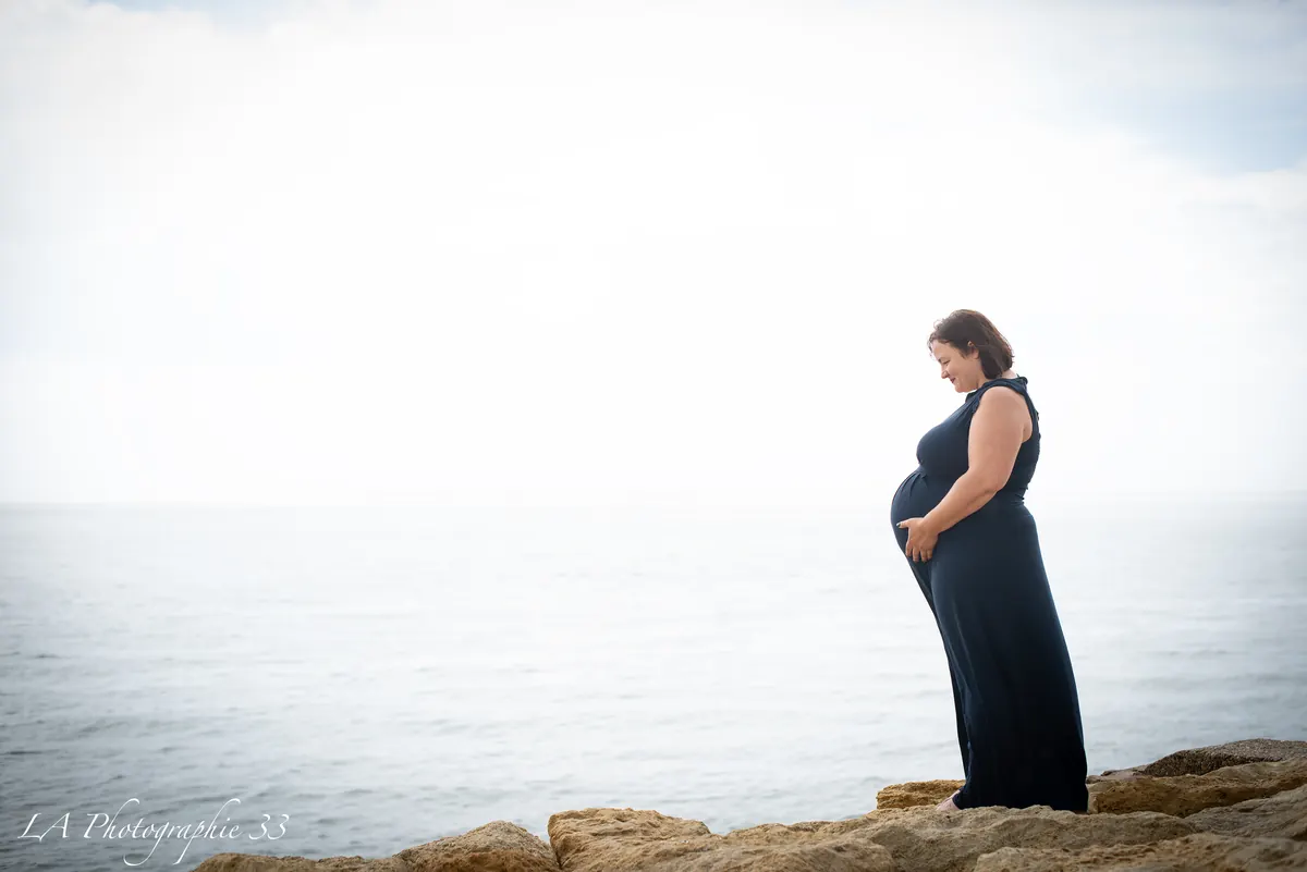 Future maman de profil face à la mer, mains posées sur son ventre, sur des rochers