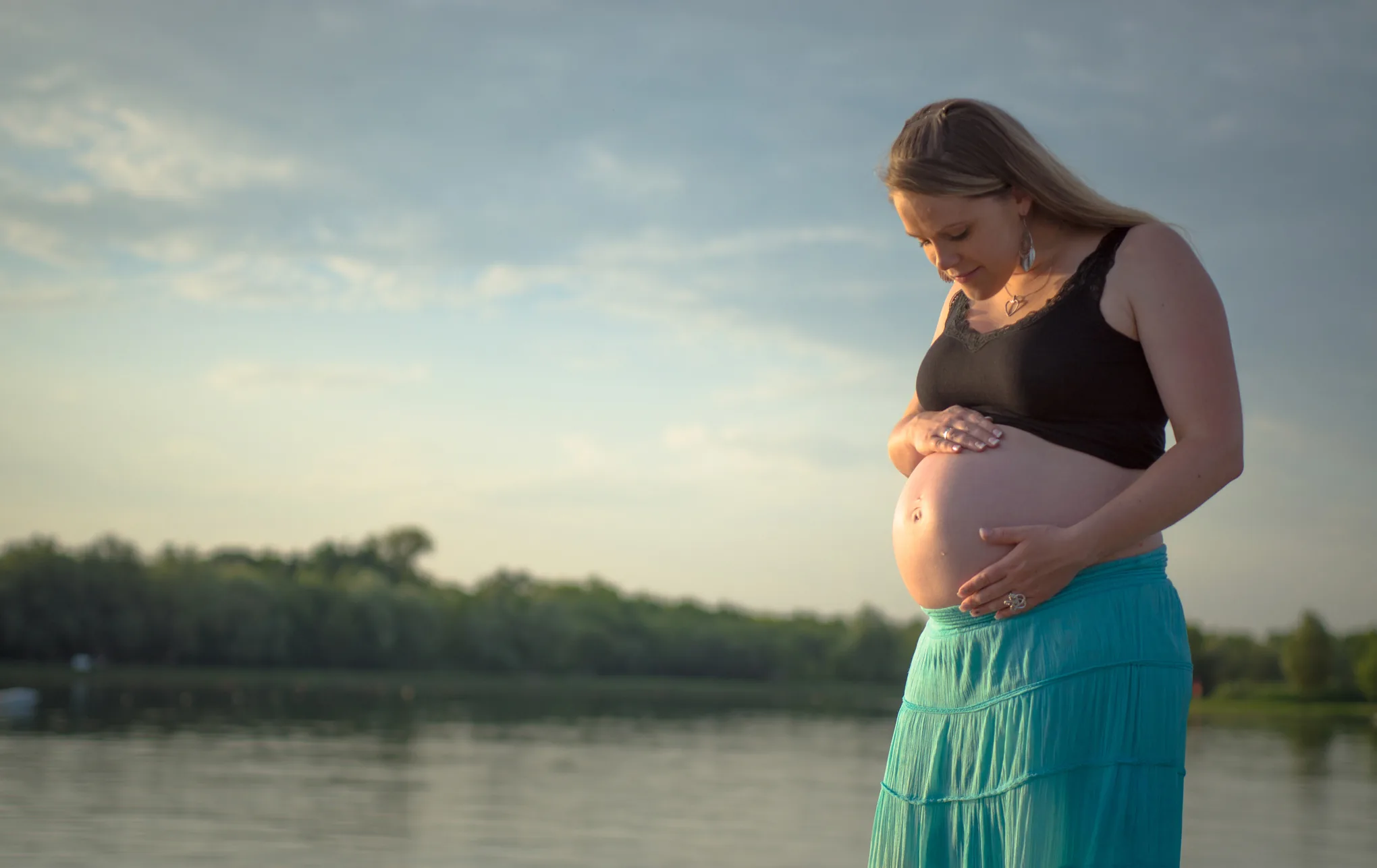 Femme enceinte de profil tenant son ventre au bord d'un lac, jupe turquoise, lumière dorée du soir